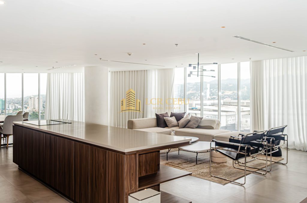 A wide-angle view of a modern, minimalist penthouse living room in Cebu IT Park featuring floor-to-ceiling windows, white drapes, a large kitchen island with wood cabinetry, and designer furniture overlooking the city and mountains.