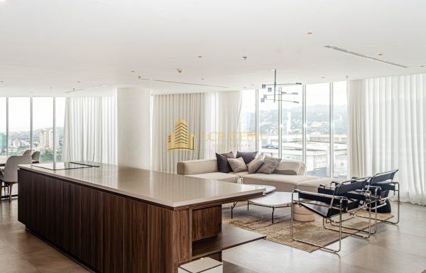 A wide-angle view of a modern, minimalist penthouse living room in Cebu IT Park featuring floor-to-ceiling windows, white drapes, a large kitchen island with wood cabinetry, and designer furniture overlooking the city and mountains.